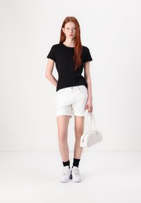 T-shirt nera a maniche corte, pantaloni di jeans bianchi, borsa bianca e sneakers bianche con calzini neri, esposti su uno sfondo semplice.