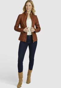 Giubbotto di pelle marrone con colletto alto, dettagli di cucitura verticale, chiusura con zip, abbinato a jeans scuri e stivali beige.