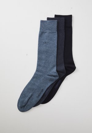 Tres pares de calcetines doblados de Calvin Klein en azul claro, gris oscuro y negro, dispuestos de manera superpuesta sobre una superficie blanca.