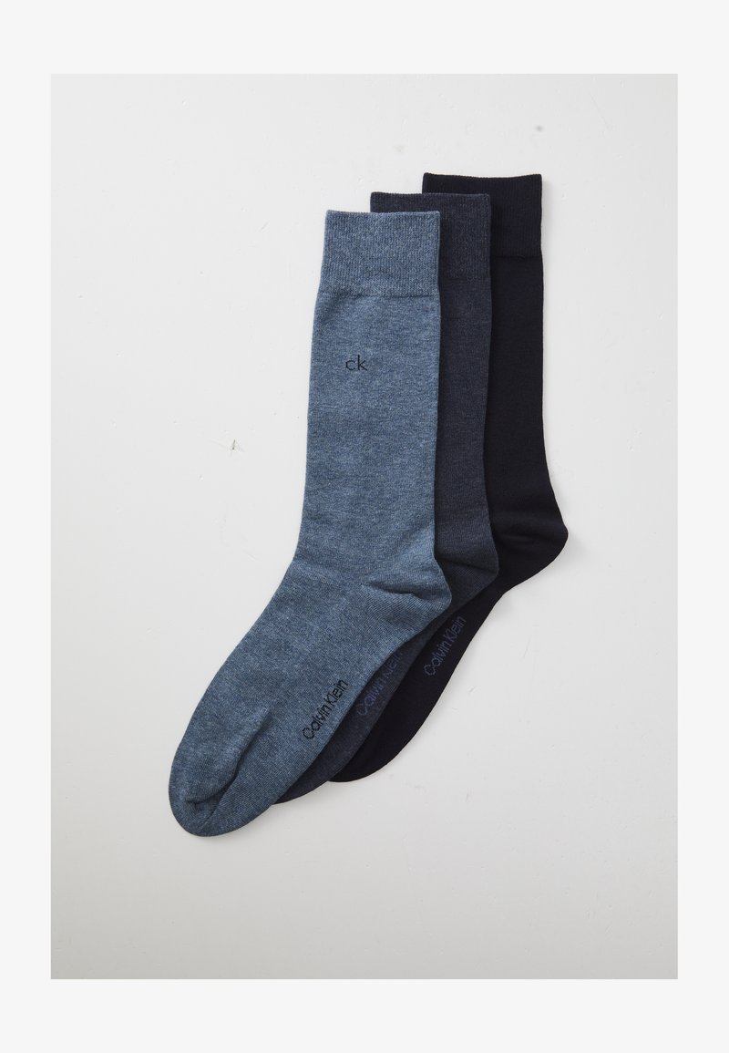 Tres pares de calcetines doblados de Calvin Klein en azul claro, gris oscuro y negro, dispuestos de manera superpuesta sobre una superficie blanca.