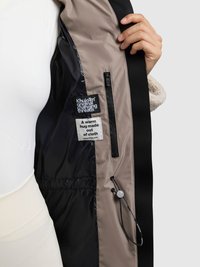 Interior de chaqueta negra y marrón con etiquetas de tela, bolsillo con cremallera y cierre de cordón. Textura suave con colores contrastantes.