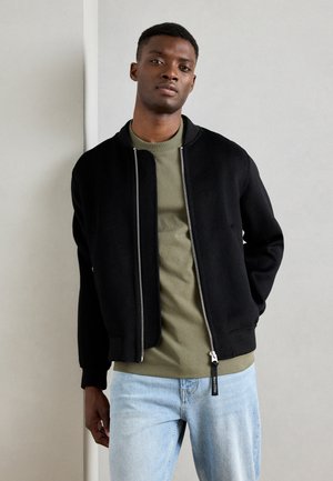 Veste bomber noire avec une texture lisse, fermeture éclair, poignets côtelés et coupe décontractée, portée sur une chemise verte olive et un jean bleu clair.