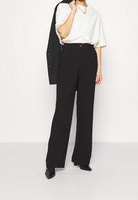 Svarta vida byxor med hög midja, med knäppning fram. I kombination med en lös vit t-shirt och en svart blazer över axeln.