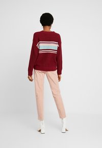 Pull côtelé couleur bordeaux avec des rayures multicolores dans le dos, accompagné d'un pantalon slim rose clair et de bottines blanches.