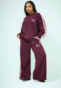 Vrouw staat met handen in de zakken, draagt wijnrode broek met wijde pijpen en bijpassende trui met "NYC 1987" en lichtroze strepen aan de zijkant.