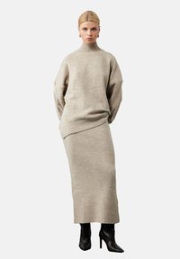 Beżowy sweter dzianinowy z wysokim kołnierzem, oversized rękawami oraz pasującą spódnicą z prążkowanej tkaniny. Modelka nosi czarne botki na obcasie. Minimalistyczny design.