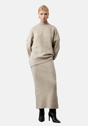 Beżowy sweter dzianinowy z wysokim kołnierzem, oversized rękawami oraz pasującą spódnicą z prążkowanej tkaniny. Modelka nosi czarne botki na obcasie. Minimalistyczny design.