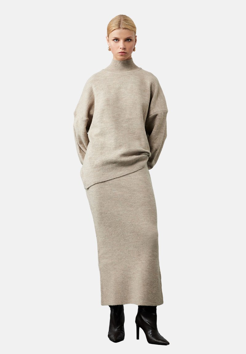Beżowy sweter dzianinowy z wysokim kołnierzem, oversized rękawami oraz pasującą spódnicą z prążkowanej tkaniny. Modelka nosi czarne botki na obcasie. Minimalistyczny design.