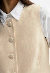 Beige linneväst med fem stora, runda knappar; bärs över en vit skjorta med krage. Har en slät textur och skräddarsydd design.