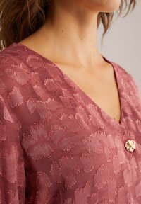 Röd texturerad blus med V-ringning och en dekorativ knapp fram. Tyget har ett subtilt djurmönster.