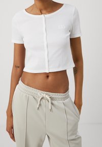 Vit ribbad croptop med korta ärmar och knäppning framtill, med en subtil broderad logotyp på vänster bröst.