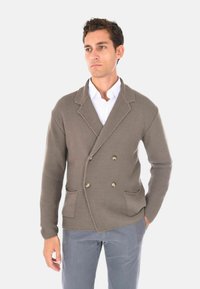 Cardigan marrone lavorato a maglia con un design doppiopetto, caratterizzato da due tasche frontali e bottoni in tonalità oro, indossato sopra una camicia bianca.