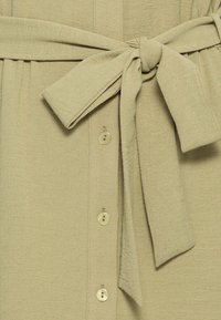 Vestido verde claro com um tecido texturizado, apresentando um cinto amarrado na cintura e pequenos botões na parte da frente. Design simples com um acabamento suave.