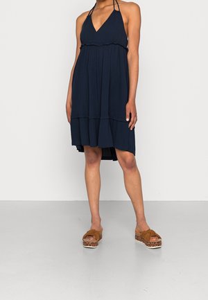 Mujer con vestido palabra de honor azul marino sin mangas con un dobladillo fruncido y sandalias de plataforma marrones con detalles tejidos.