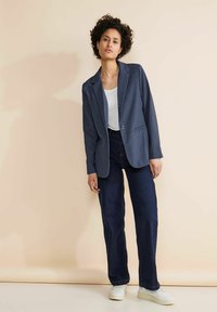 Blazer blu navy con un bottone, indossato sopra una canottiera bianca. Abbinato a jeans in denim scuro e sneakers bianche. Texture liscia.