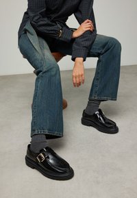 Zwarte leren schoenen met een gespsluiting, stevige zolen en een glanzende afwerking, gecombineerd met losse blauwe spijkerbroeken en grijze sokken.