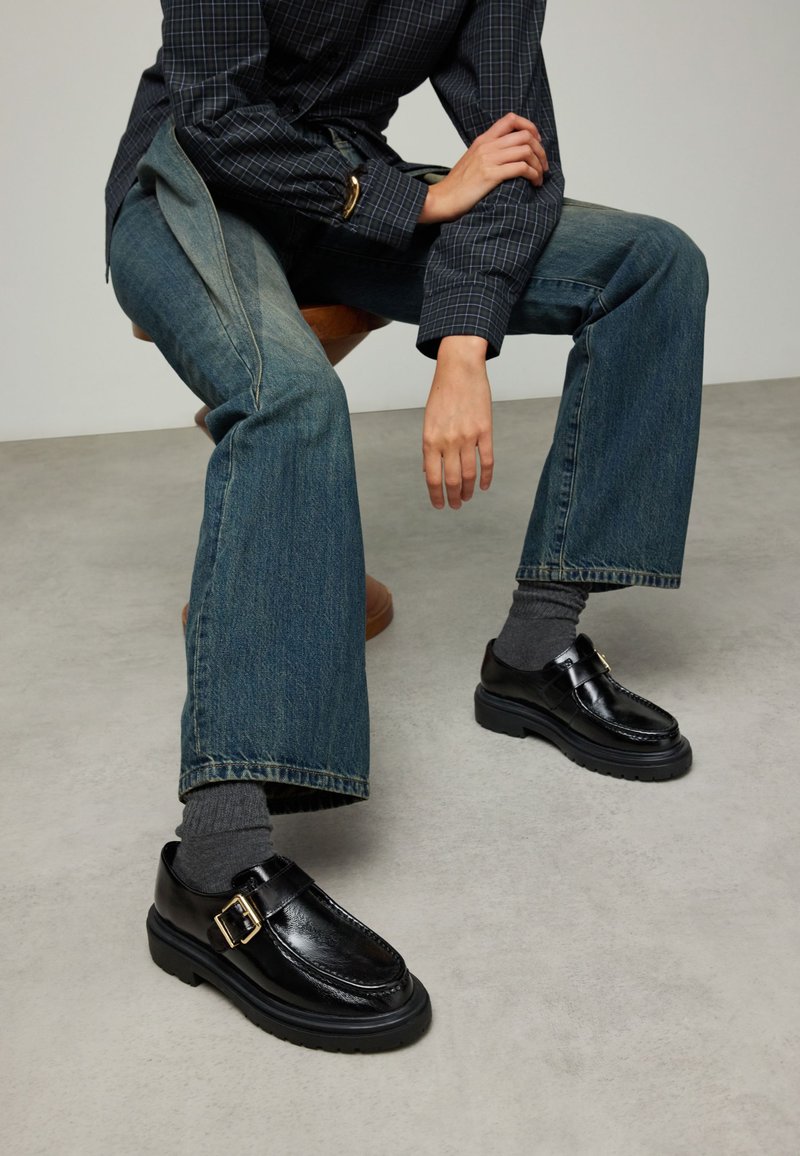 Zwarte leren schoenen met een gespsluiting, stevige zolen en een glanzende afwerking, gecombineerd met losse blauwe spijkerbroeken en grijze sokken.