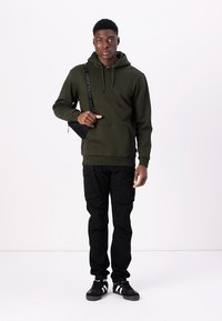 Olive green hoodie with a front pocket, black cargo pants, and black sneakers with white stripes. Crossbody bag with a logo strap.