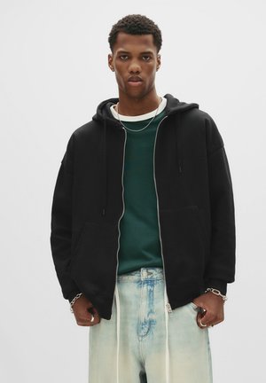 Young man wearing a black zip-up hoodie, green shirt, light blue jeans, and silver jewelry, standing against a plain white background.