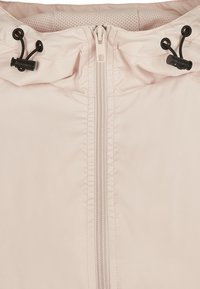 Giacca rosa chiaro con zip frontale, cordini neri regolabili sul cappuccio e fodera in rete testurizzata. Materiale liscio e resistente all'acqua.