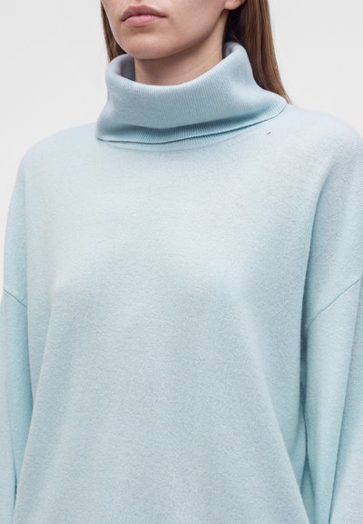 Femme portant un pull à col roulé en tissu doux bleu clair avec manches longues et col plié.