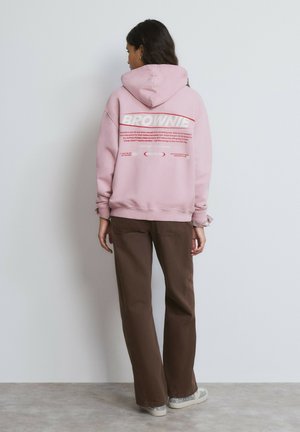 Sweat à capuche rose avec un texte graphique blanc au dos, associé à un pantalon large marron et des chaussures argentées, présenté sur un fond neutre.