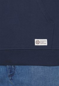 Gros plan d'un sweat-shirt bleu foncé avec poche avant et étiquette blanche indiquant "Exploration without compromise" au-dessus d'un jean en denim bleu.