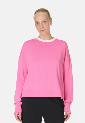 Woman with blonde hair in a pink long-sleeve sweatshirt over a white undershirt, wearing black pants, standing against a plain background.