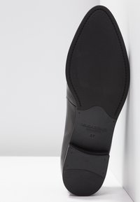 Chaussure habillée en cuir noir montrant la semelle texturée avec le nom de marque V. Sapon et la taille 37 sur fond blanc.