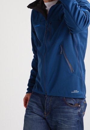 Homme portant une veste bleue Mammut GORE Windstopper avec poches zippées et jean bleu, main près du visage, cadré au niveau du cou.