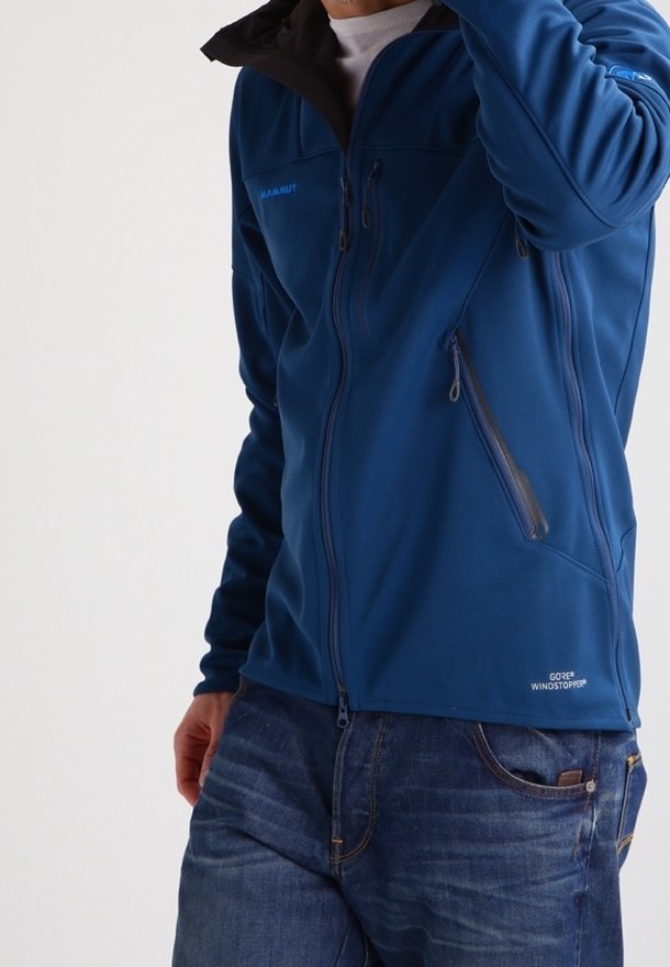 Homme portant une veste bleue Mammut GORE Windstopper avec poches zippées et jean bleu, main près du visage, cadré au niveau du cou.