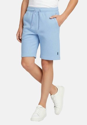 Persoon die lichtblauwe korte broek met trekkoord en zakken draagt, samen met witte sneakers, staand met één hand in de zak en de benen gekruist.