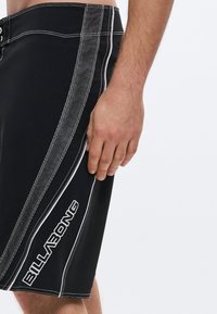 Μαύρο μαγιό σορτς με λευκά ραφτά, εναλλασσόμενες μονές και τελείες ρίγες, με το λογότυπο "BILLABONG" στο αριστερό πόδι.