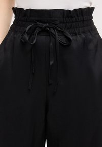 Μαύροι ψηλόμεσοι παντελόνες με ελαστική μέση που μαζεύει και δεμένο φιόγκο μπροστά, φορεμένοι με λευκό τοπ.