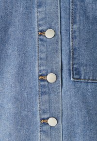 Denim jurk van lichtblauwe stof, vijf metalen knopen met gegraveerde tekst en een borstzak met een effen rand.