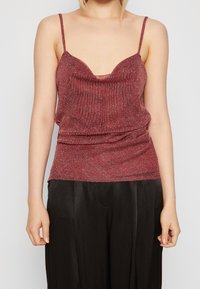 Donna che indossa un top canotta senza maniche color bordeaux glitterato abbinato a pantaloni neri in satin larghi, su uno sfondo neutro.