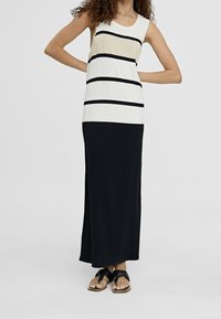 Donna che indossa un vestito lungo senza maniche bianco e nero con righe orizzontali, abbinato a sandali neri con zeppa.