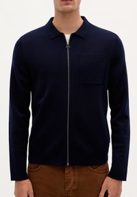 Marineblå zippered sweater med krave, lange ærmer og en brystlomme, lavet af et glat strikstof. Har en sølvfarvet lynlås.