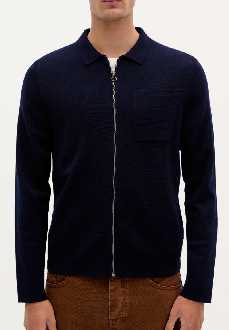 Marineblå zippered sweater med krave, lange ærmer og en brystlomme, lavet af et glat strikstof. Har en sølvfarvet lynlås.