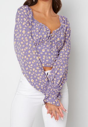 Femme portant un crop top manches longues lavande avec un imprimé floral jaune et blanc et un pantalon blanc taille haute, les mains croisées devant.