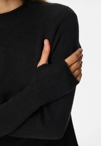 Osoba nosiaca čierny sveter s dlhým rukávom s otvormi na palce, skrížené ruky, pravou rukou drží ľavý horný rameno.