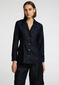 Camicia in denim scuro a maniche lunghe, colletto a punta, chiusura frontale con bottoni e un leggero lucido. Indossata con pantaloni corti scuri abbinati.