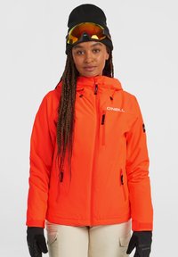 Heldere oranje skijas met een rits aan de voorkant, verstelbare capuchon en meerdere zakken, gecombineerd met zwarte handschoenen en een zwarte beanie.
