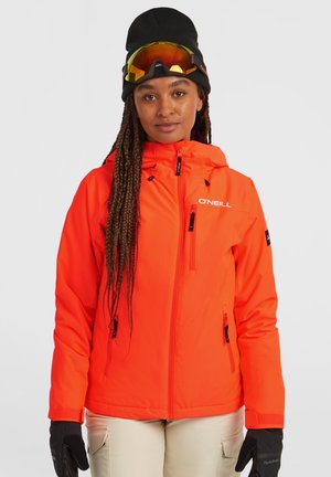 Heldere oranje skijas met een rits aan de voorkant, verstelbare capuchon en meerdere zakken, gecombineerd met zwarte handschoenen en een zwarte beanie.