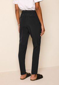 Pantalon noir coupe slim avec ceinture élastique, doté de deux poches arrière et d'une texture lisse, porté avec des sandales noires.