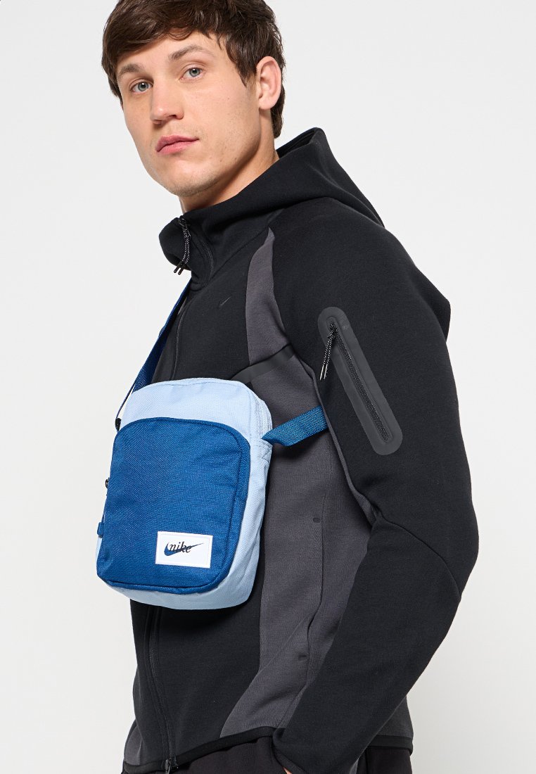 Bolso bandolera azul y azul claro hecho de tela texturizada, con un bolsillo delantero y una correa ajustable, llevado sobre una chaqueta negra.