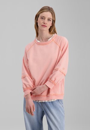 Frau mit schulterlangem Haar, die einen lockeren pinkfarbenen Sweatshirt und hellblaue Jeans vor einem einfarbigen grauen Hintergrund trägt.