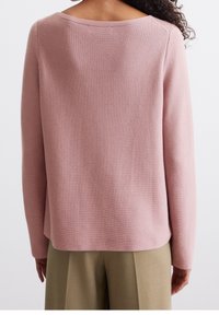 Personne aux cheveux bouclés portant un pull rose clair en tricot et un pantalon beige, vue de dos sur un fond uni.