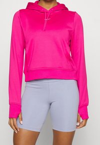 Femme portant un hoodie rose vif et un short de cyclisme ajusté gris clair, montrée du cou à mi-cuisse, se tenant devant un fond uni.