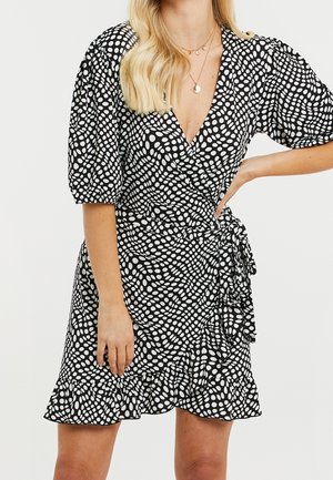 Kobieta w czarnej sukience kopertowej w nieregularne białe grochy, z bufkami na rękawach, falbaną na dole i warstwowymi złotymi naszyjnikami.